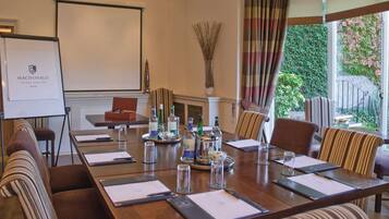 A conference room with a long table, chairs, a projector screen, and a view of greenery outside.