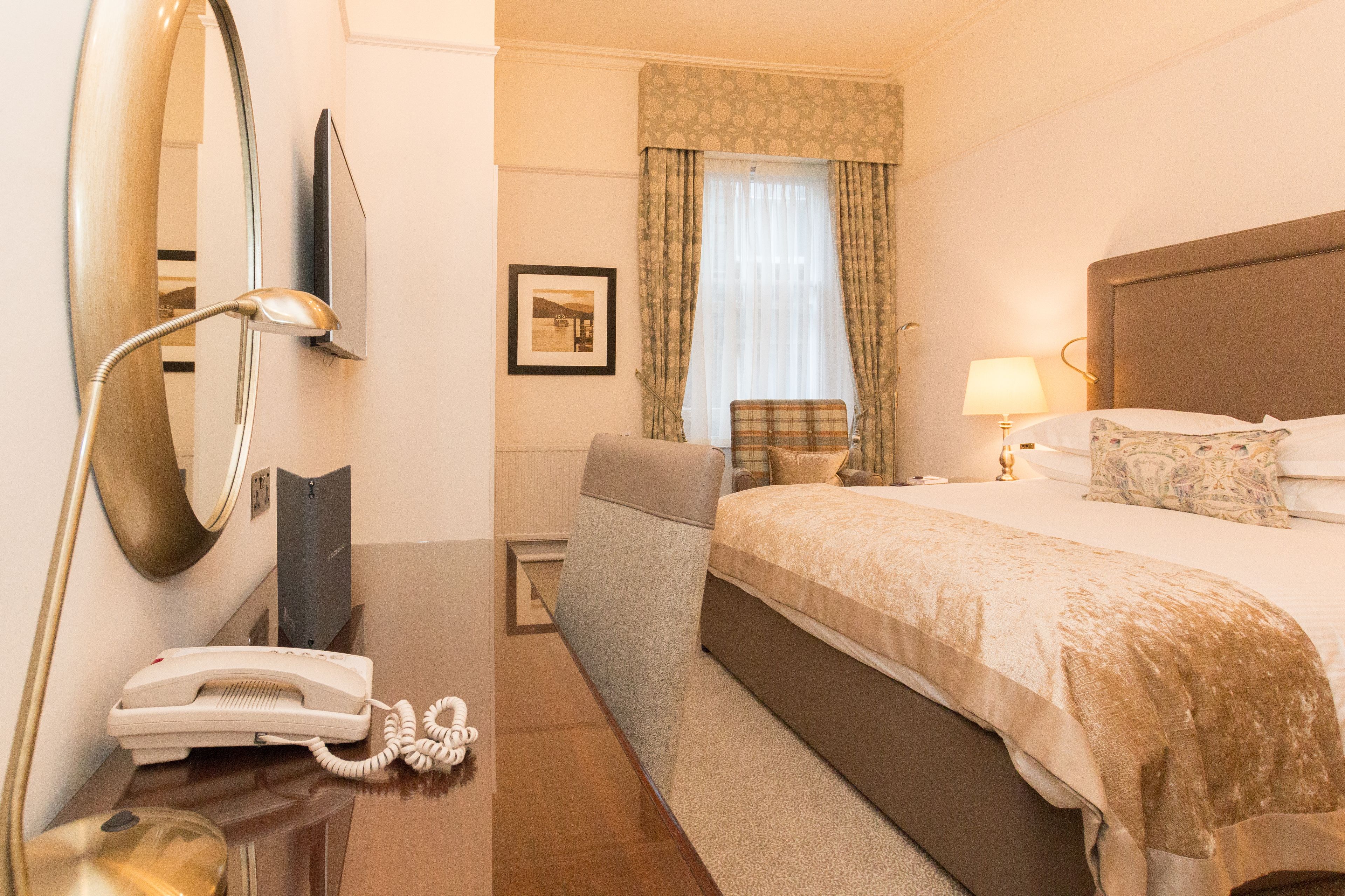 A hotel room with a bed, a desk with a telephone, a chair, a lamp, and a window with curtains.