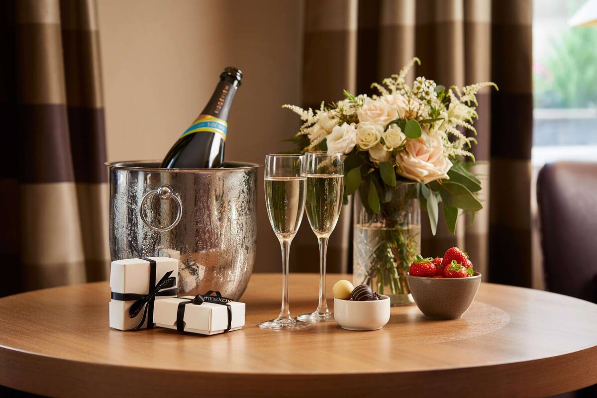 A table with a champagne bottle, two glasses, a bowl of strawberries, and a bouquet of flowers.