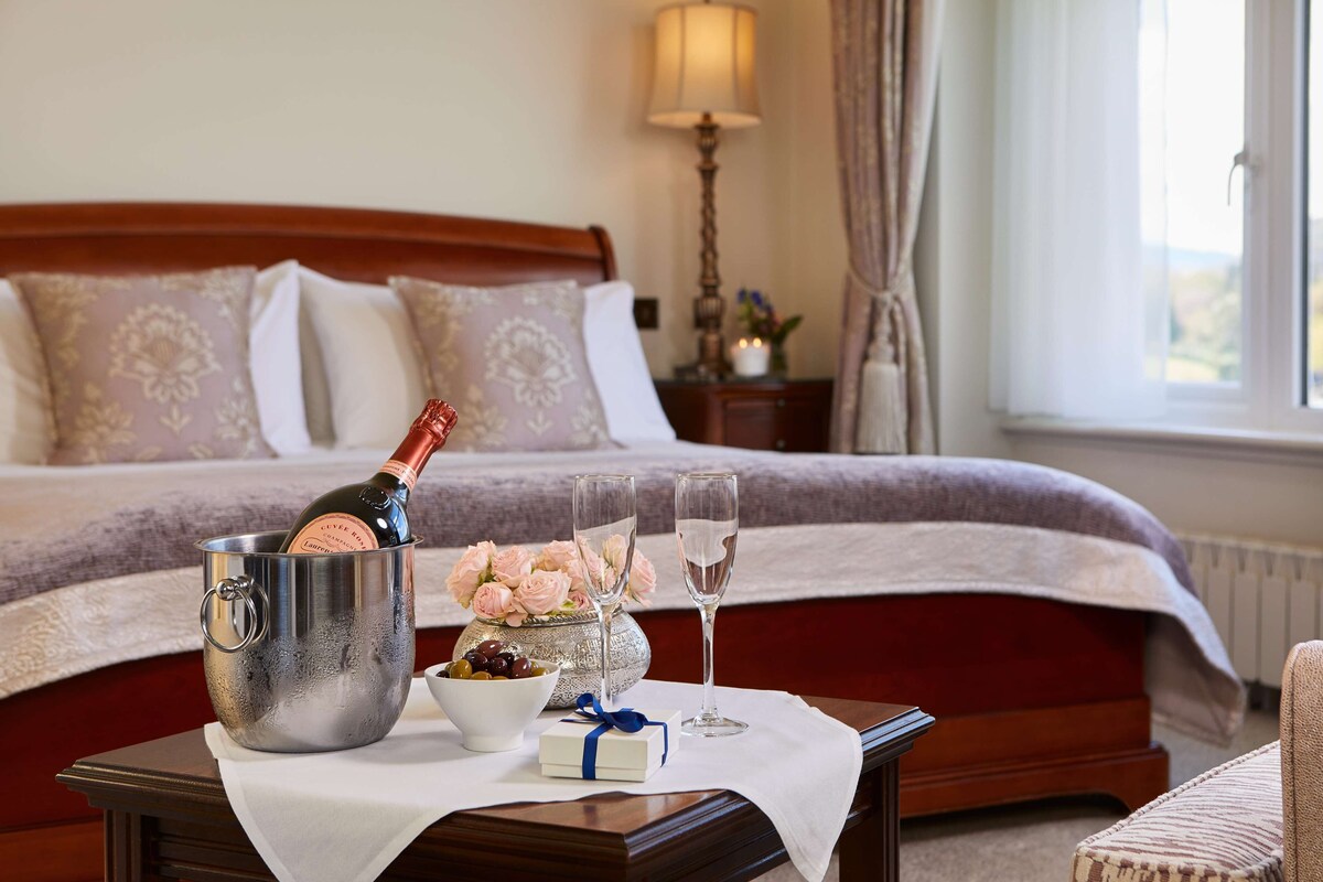 A hotel room with a bed, a bedside table with a bottle of champagne, two glasses, a bowl of olives, and a small gift box.