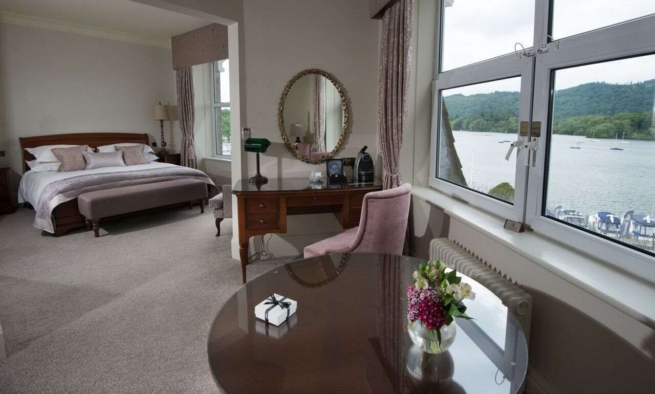 A hotel room with a bed, a desk, a chair, a round table with a vase of flowers, a mirror, and a view of a lake through the window.