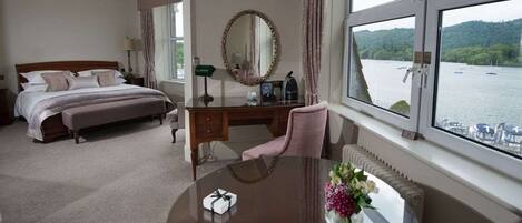 A hotel room with a bed, a desk, a chair, a round table with a vase of flowers, a mirror, and a view of a lake through the window.