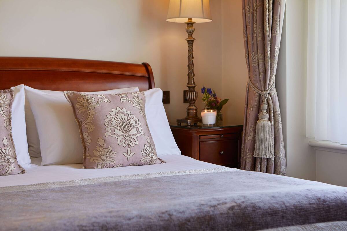 A neatly made bed with floral pillows, a bedside table with a lamp, and a vase with flowers.