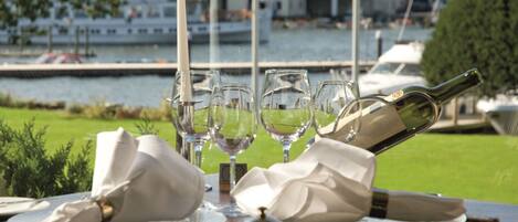 A table set for dining with wine glasses, a bottle, and a bell, overlooking a marina with boats and greenery.