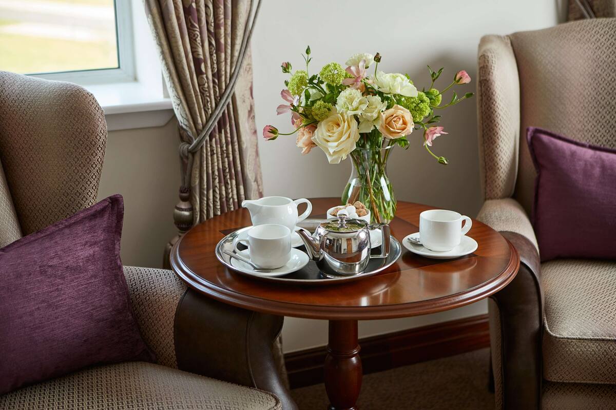 A round wooden table with a tea set, a vase of flowers, and two chairs with purple cushions.