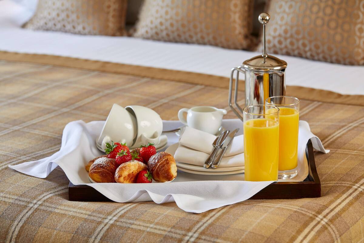 A breakfast tray with croissants, strawberries, a coffee pot, cups, and glasses of orange juice on a checkered tablecloth.