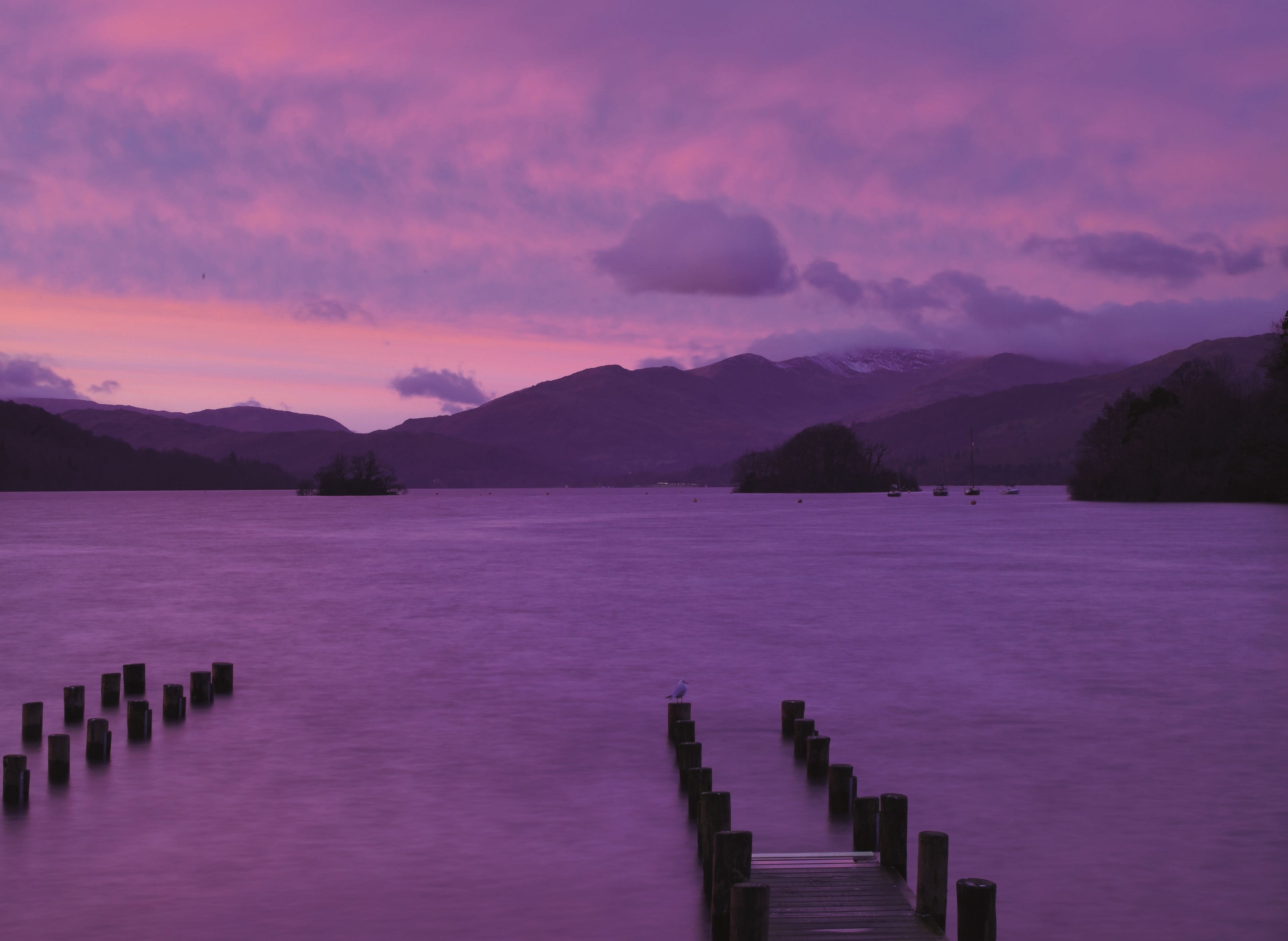 A calm lake with a wooden dock extending into the water, surrounded by mountains and a purple sky.