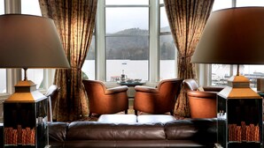 A room with a leather sofa, two armchairs, a glass coffee table, and two lamps. A large window offers a view of a lake and boats. - Macdonald Old England Hotel & Spa (Windermere)