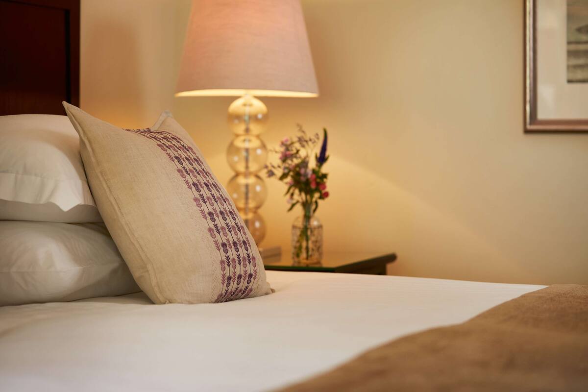 A neatly made bed with white and beige pillows, a decorative lamp, and a vase with flowers on a bedside table.