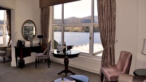 A room with a large window overlooking a lake, a round table with a vase and bottles, a chair, and a desk with a mirror and lamp. - Macdonald Old England Hotel & Spa (Windermere)