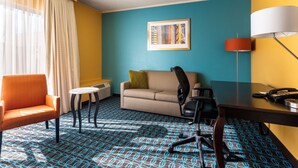 A hotel room with a blue patterned carpet, a beige sofa, an orange armchair, a small round table, a desk with a lamp, and a framed picture on the wall. -  Chester Inn & Suites (Sharonville)