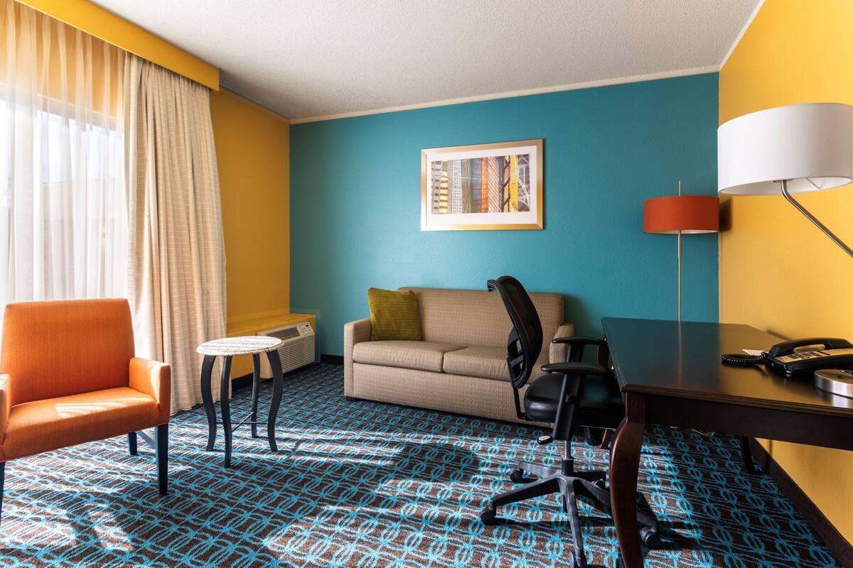 A hotel room with a blue patterned carpet, a beige sofa, an orange armchair, a small round table, a desk with a lamp, and a framed picture on the wall.