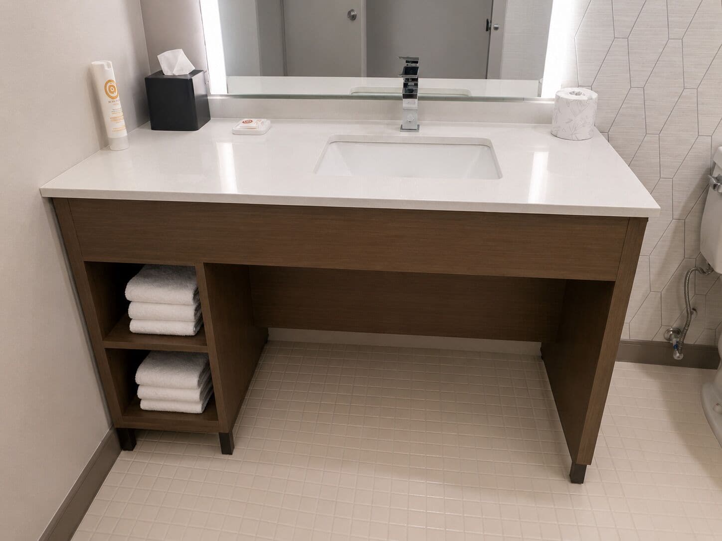 A bathroom with a white sink, a mirror above it, a towel rack with folded towels, and a toilet paper roll on the counter.