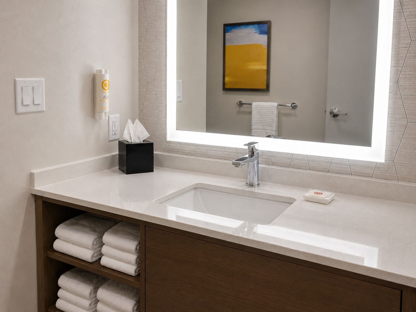 A hotel bathroom with a large mirror, a sink, a towel rack, and a shelf with folded towels.