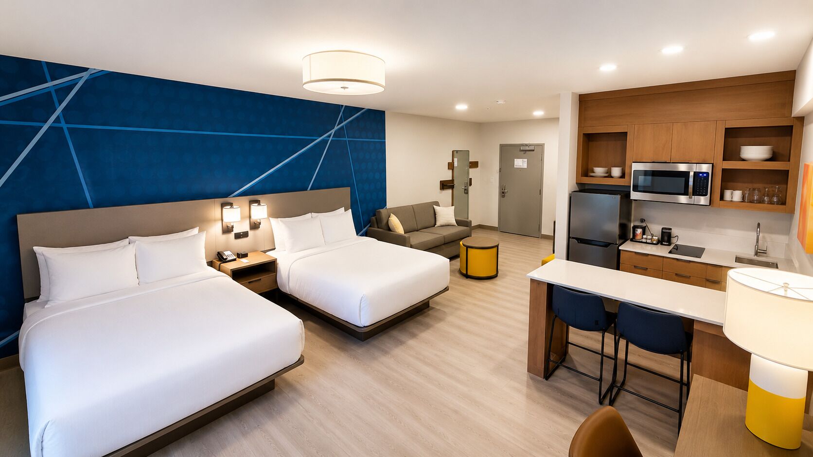 A hotel room with two beds, a sofa, a desk with a chair, a kitchenette, and a bathroom.