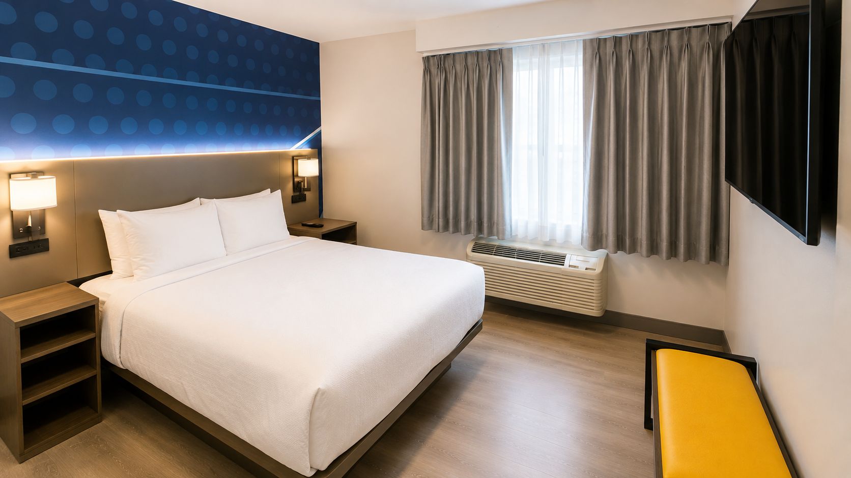 A hotel room with a bed, bedside tables, a heater, a window with curtains, and a yellow bench.