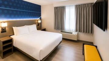 A hotel room with a bed, bedside tables, a heater, a window with curtains, and a yellow bench.