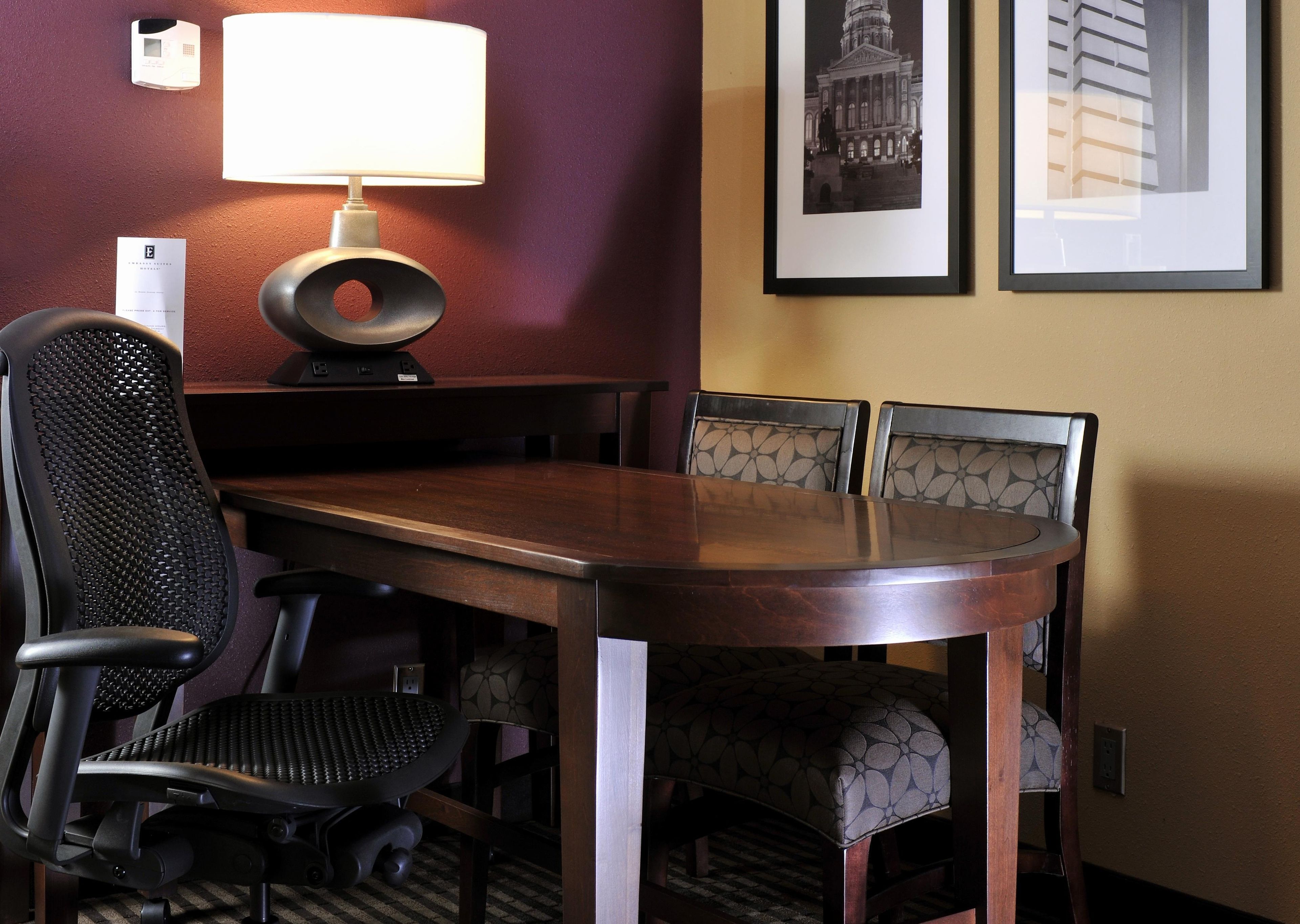 A conference room with a wooden table, four chairs, a standing lamp, and two framed pictures on the wall.