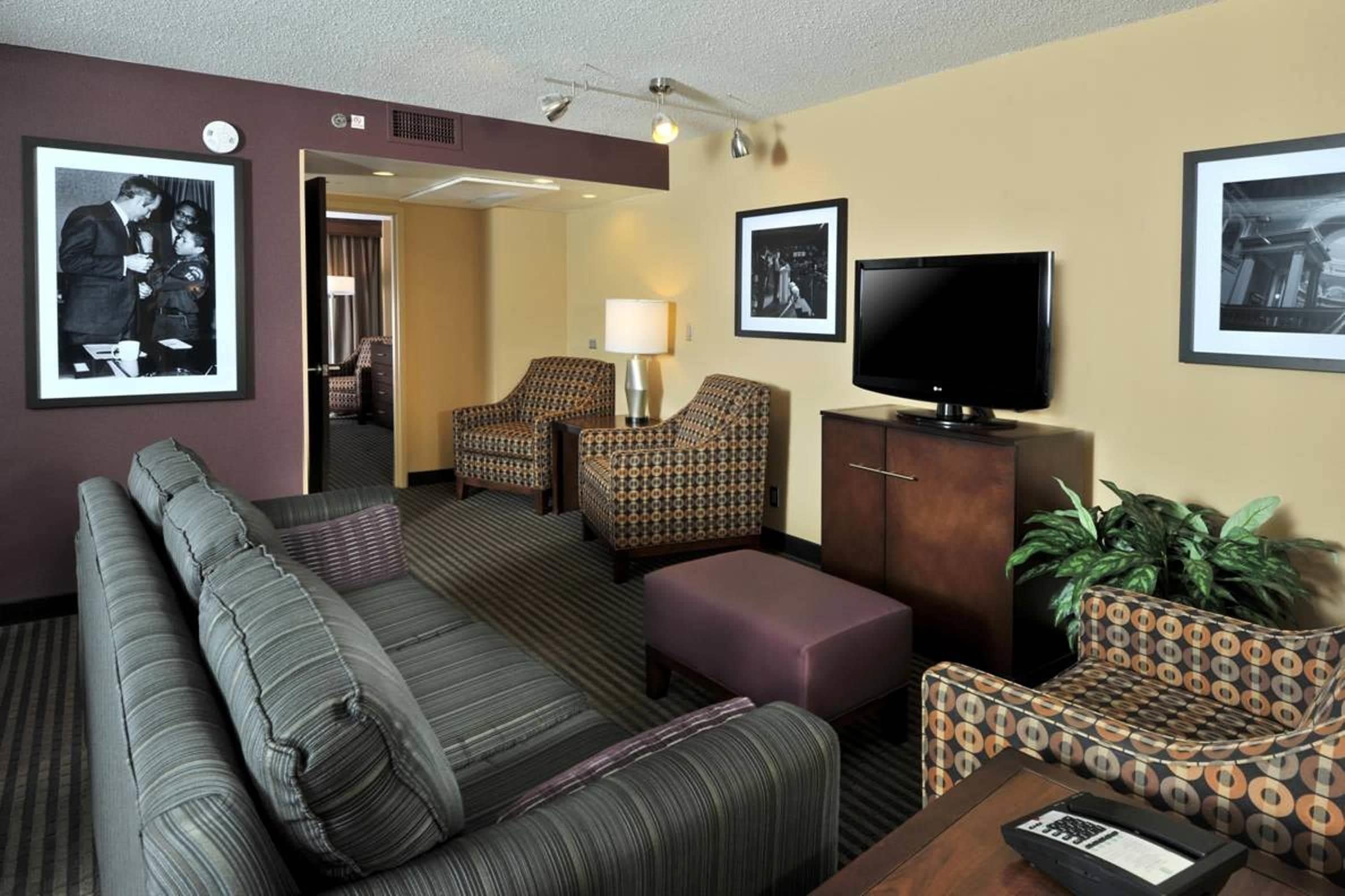 A hotel room with a sofa, armchair, ottoman, television, and framed pictures on the walls.