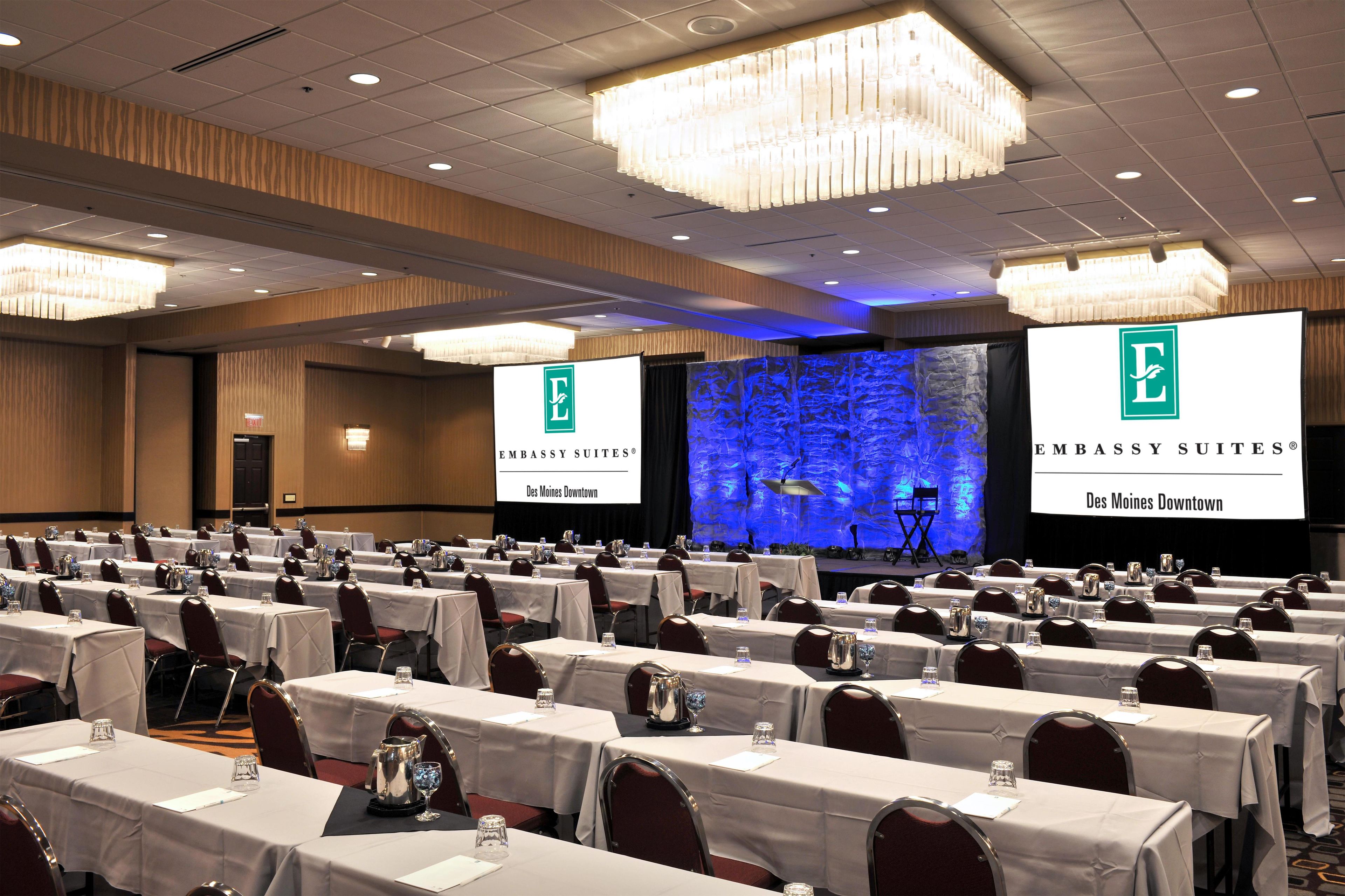 Une salle de conférence avec des rangées de tables, des chaises et une scène sur laquelle est affiché le logo de l’hôtel Embassy Suites Des Moines Downtown.