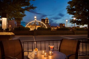Un área de comedor al aire libre con una mesa dispuesta para dos personas, un horizonte urbano de fondo y una noria iluminada contra el cielo vespertino.