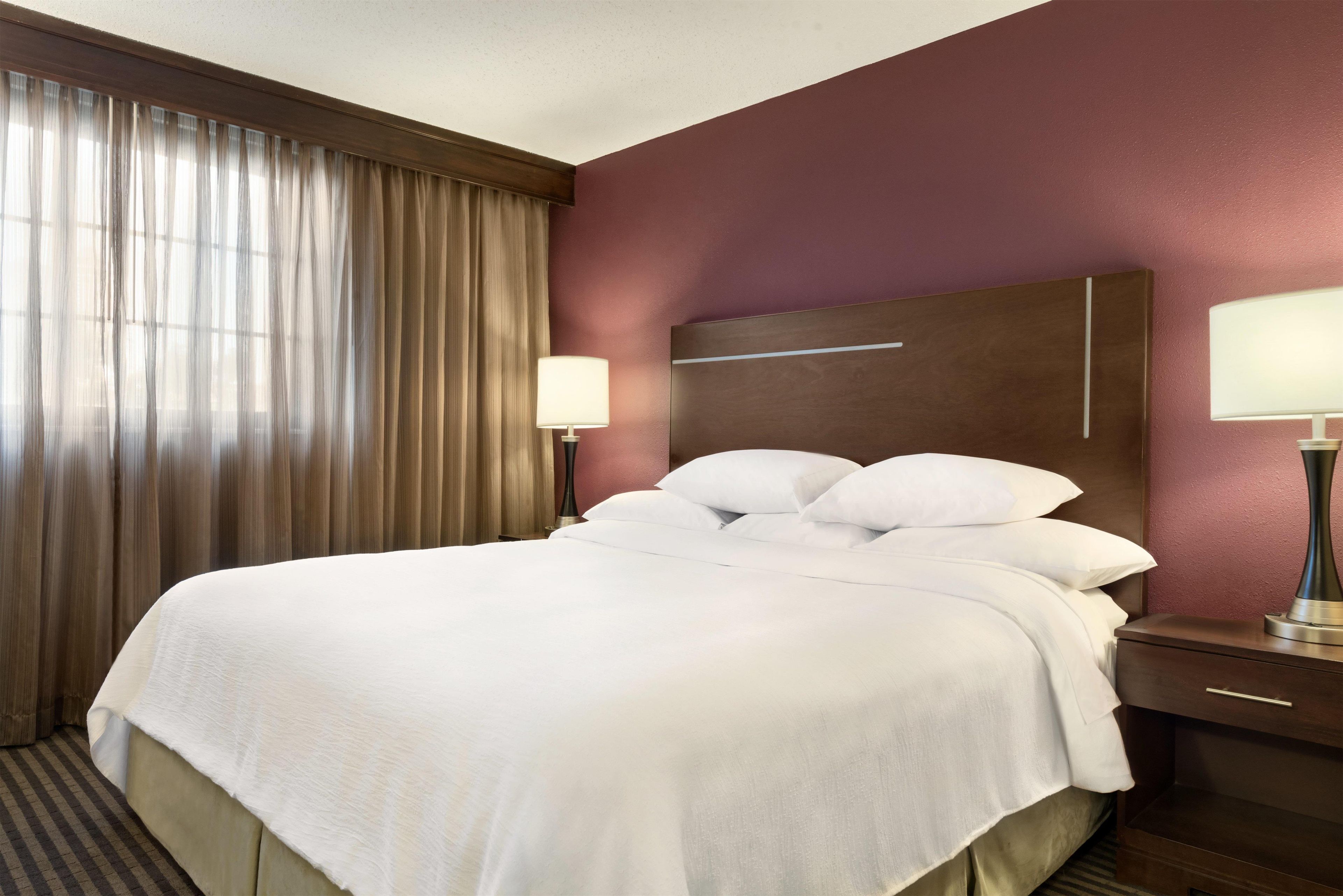 A hotel room with a bed, curtains, a nightstand, a lamp, and a wall with a textured finish.