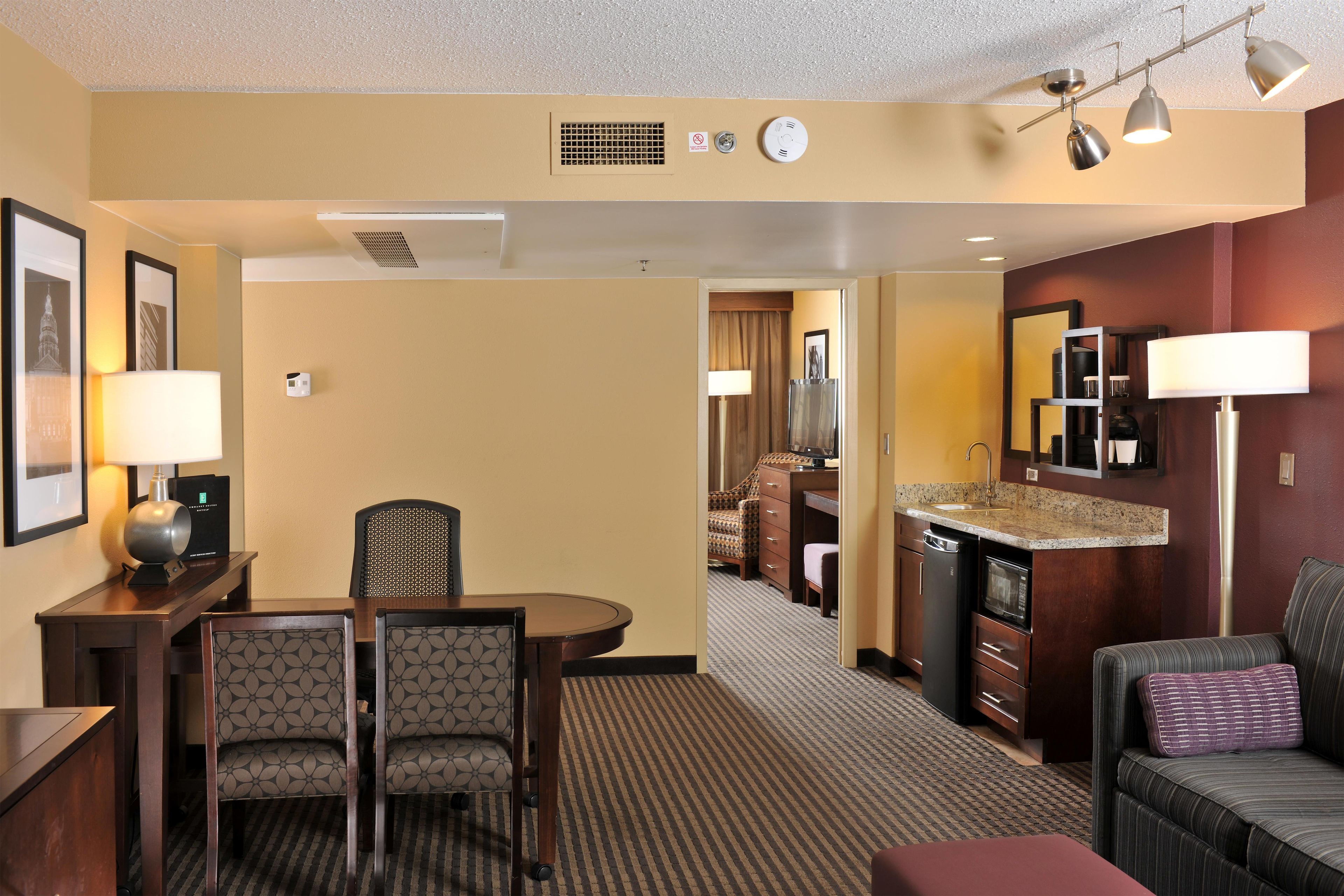 A hotel room with a dining area, a kitchenette, a living area with a sofa, and a hallway leading to a bathroom.