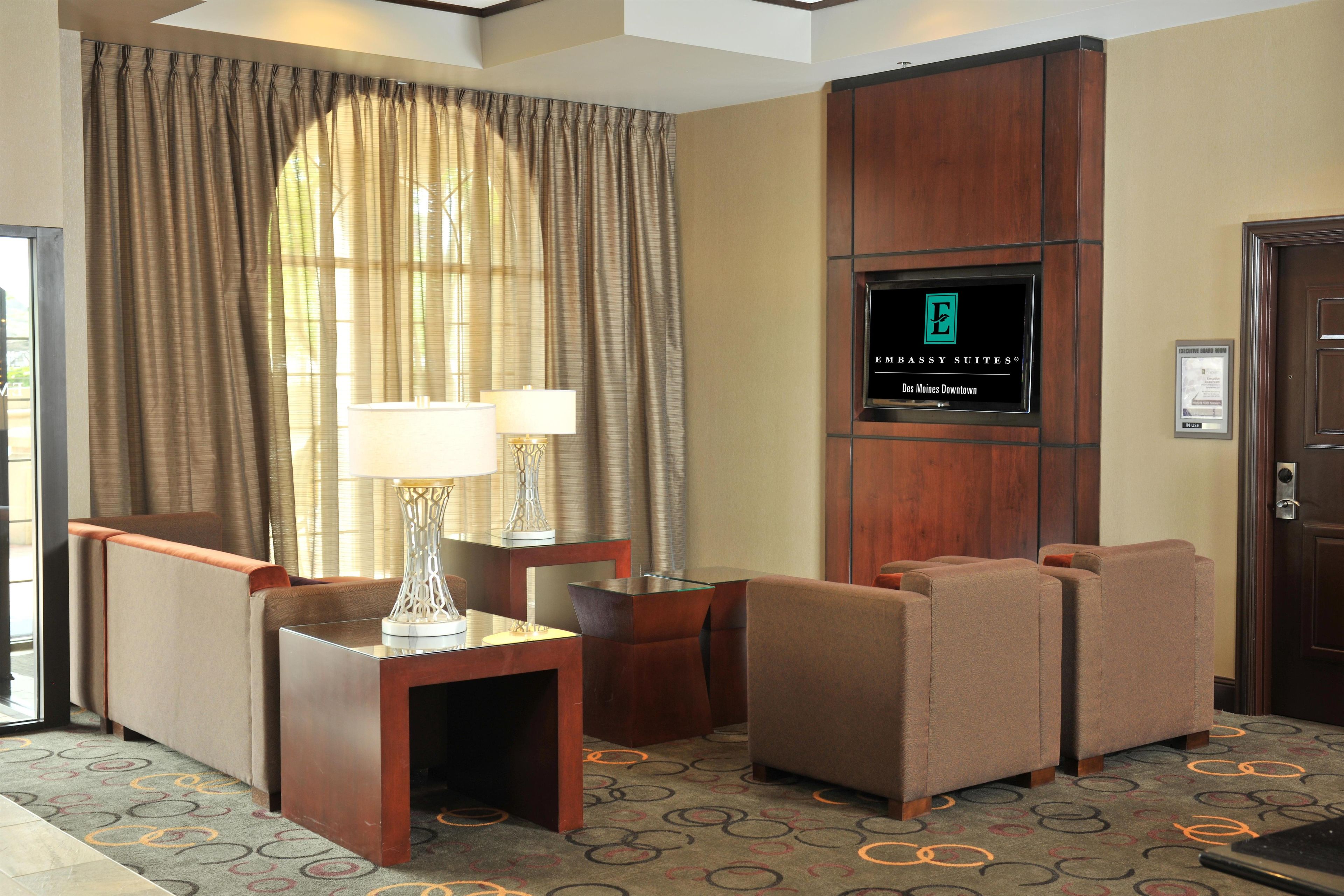 Habitación de hotel con televisor montado en la pared, una zona de estar con dos sillones y una mesita, y un ventanal con cortinas.