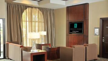 Habitación de hotel con televisor montado en la pared, una zona de estar con dos sillones y una mesita, y un ventanal con cortinas.