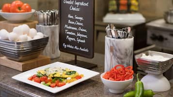 Un bufé de desayuno con un cartel que enumera los ingredientes para los omelettes, tazones de huevos, tomates y pimientos, y una jarra de mármol con cubiertos.