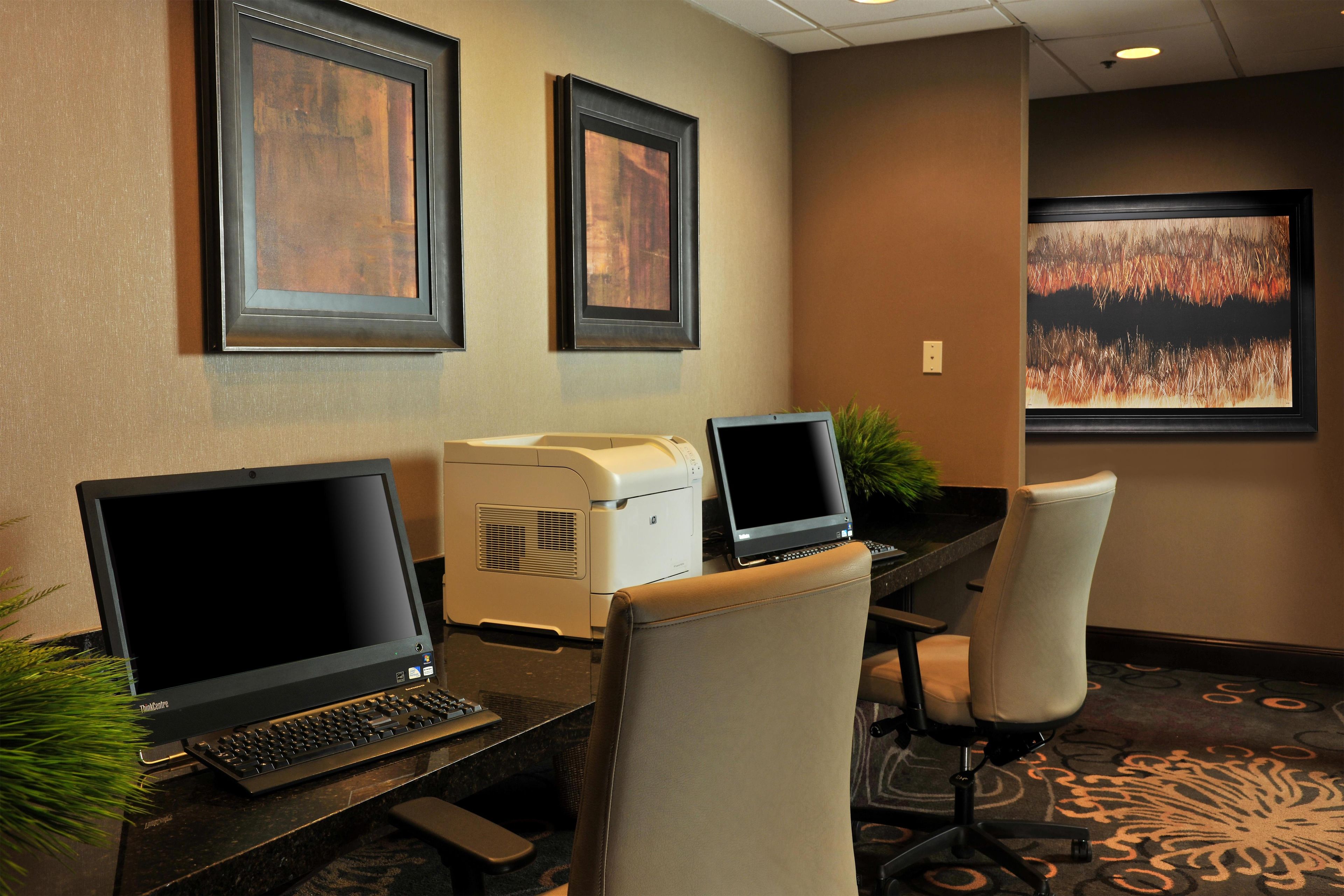 Een hotelkamer met een bureau, twee laptops, een printer en een groot raam met uitzicht op een bos.