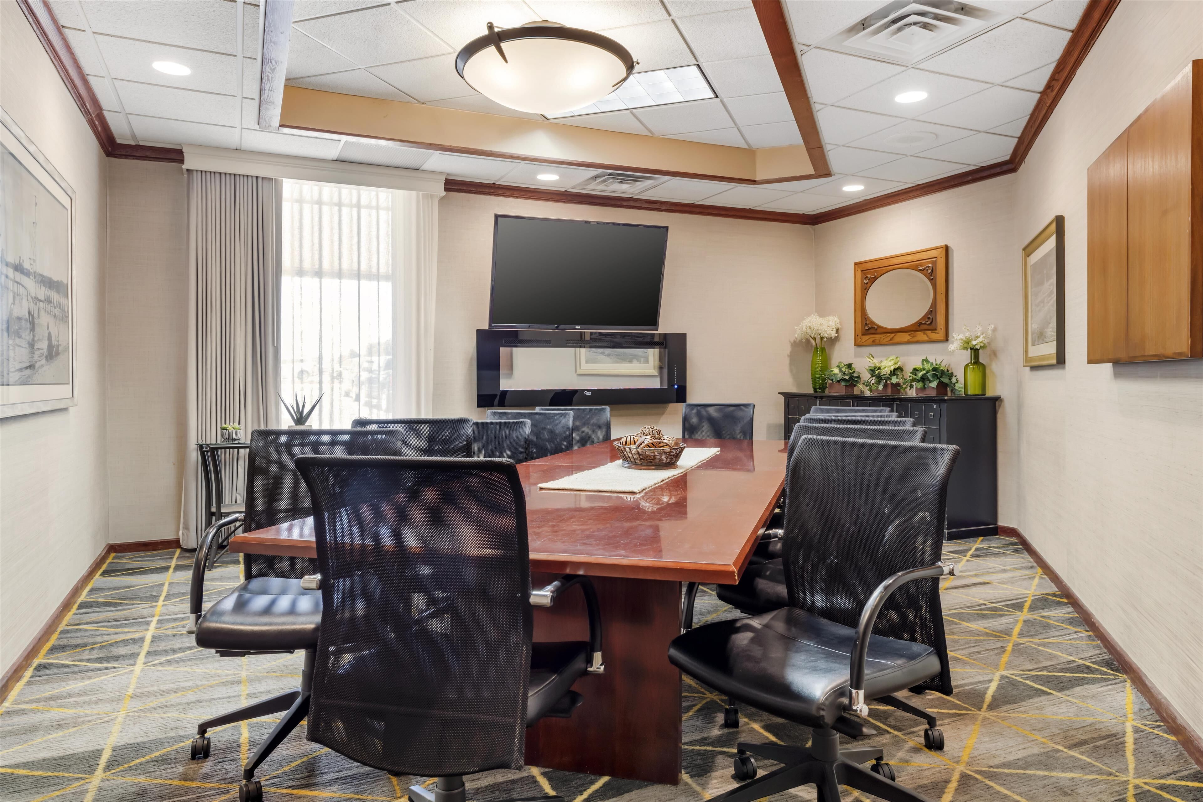 A conference room with a large table, black chairs, a flat-screen TV, and a decorative vase with flowers.