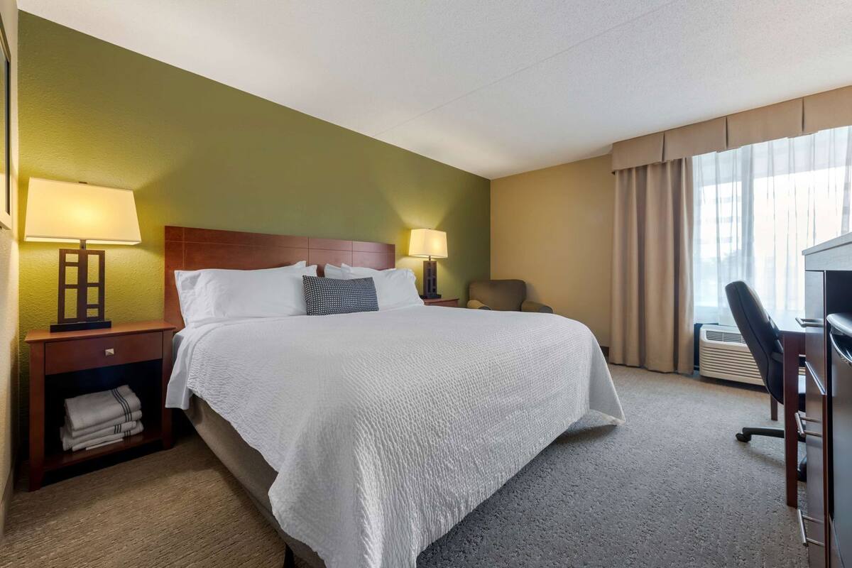 A hotel room with a bed, two bedside lamps, a chair, a desk, and a window with curtains.