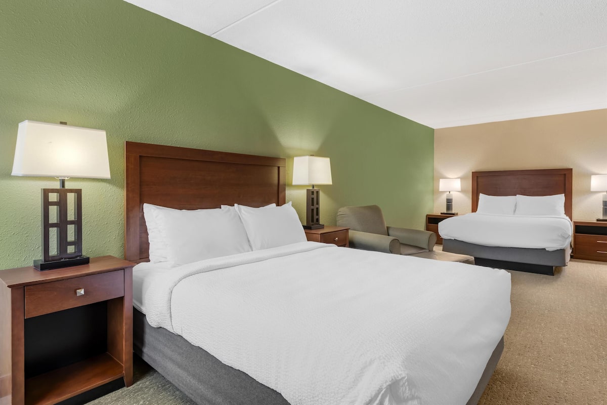 A hotel room with two beds, a wooden headboard, a nightstand with a lamp, a chair, and a carpeted floor.