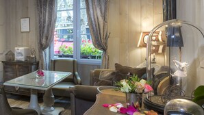 Una habitación acogedora con una mesa de vidrio, asientos acolchados, un jarrón con flores, una lámpara y una ventana con cortinas.