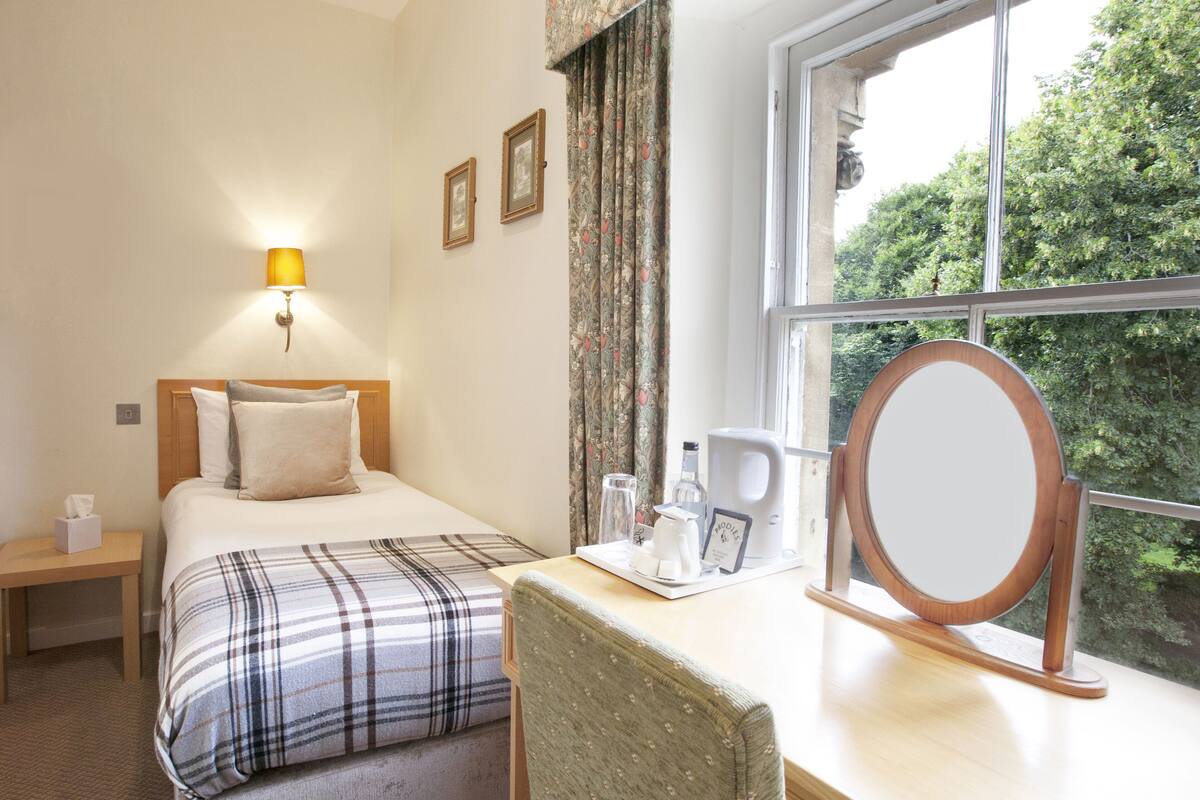 A hotel room with a bed, bedside table, lamp, window with curtains, and a desk with a mirror and coffee maker.
