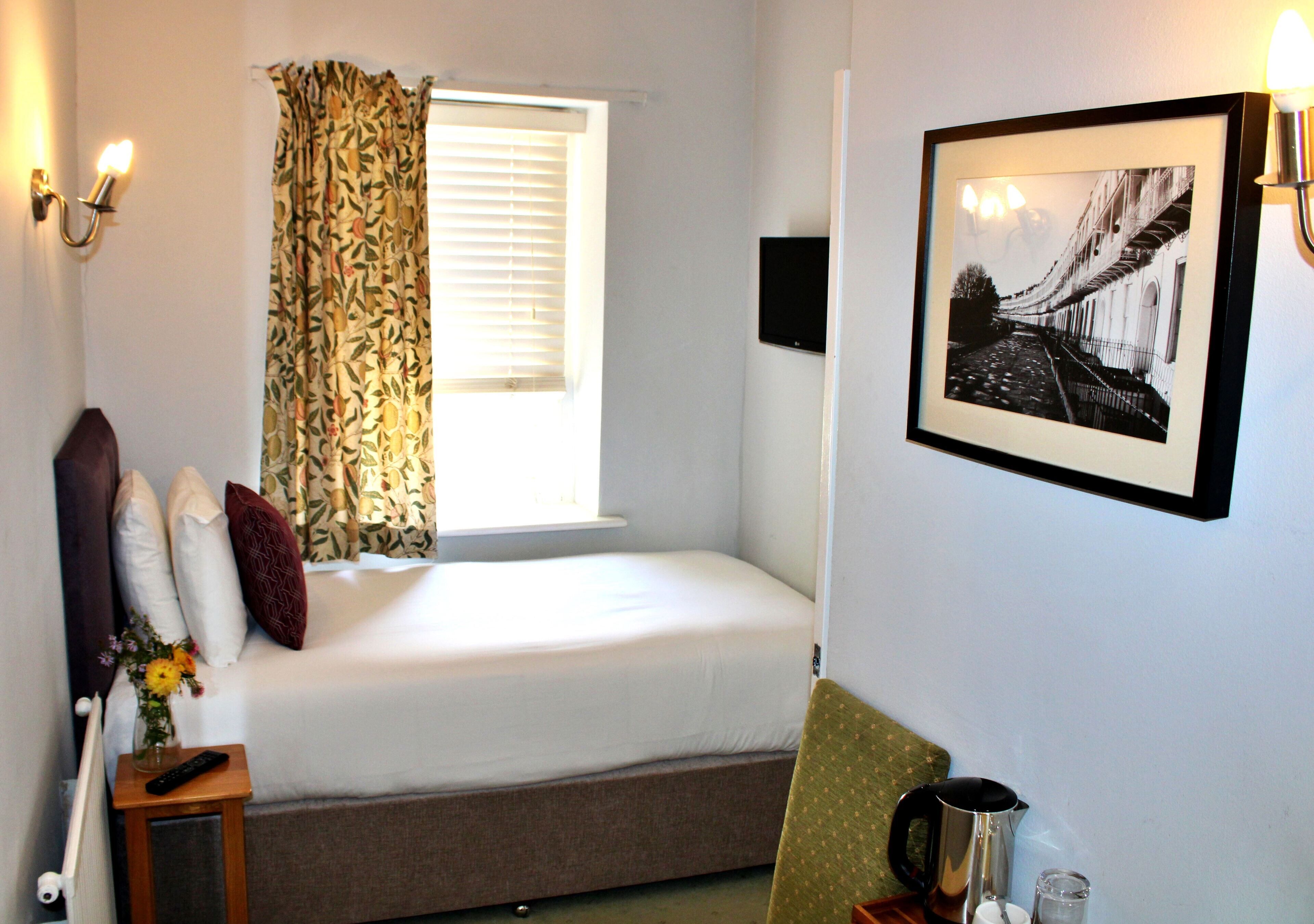 A small, neatly arranged bedroom with a single bed, a small table with a remote control, a framed picture on the wall, and a window with curtains.