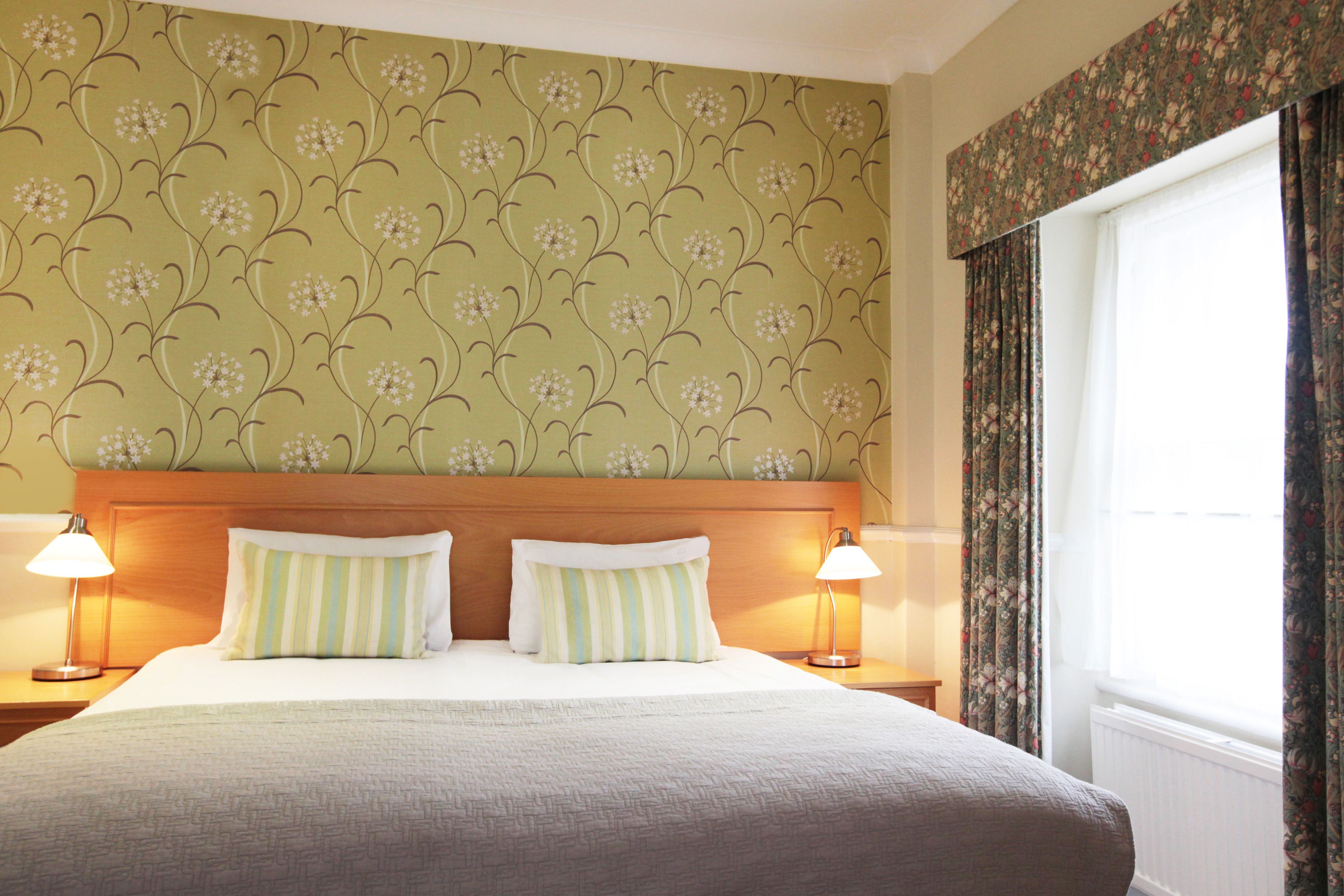 Habitación de hotel con una cama, dos lámparas de noche, papel tapiz floral y una ventana con cortinas.