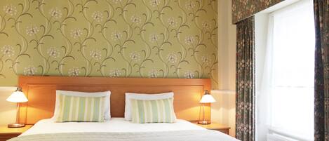 A hotel room with a bed, two bedside lamps, floral wallpaper, and a window with curtains.