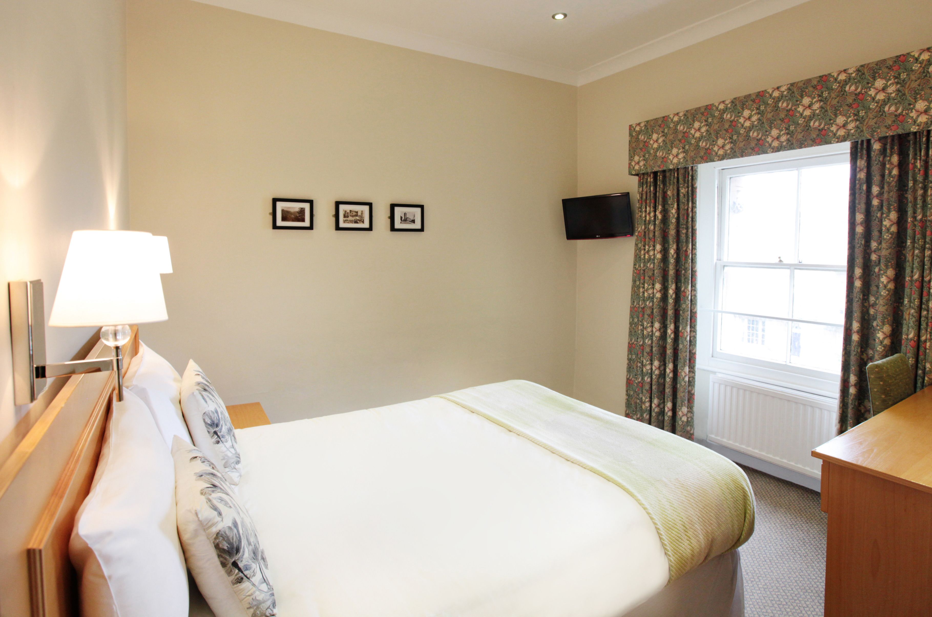 A hotel room with a bed, a desk, a lamp, a TV, and three framed pictures on the wall.
