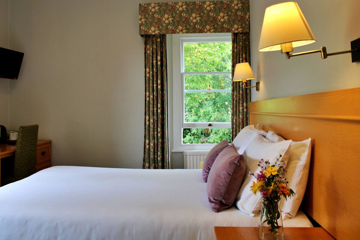 A hotel room with a bed, a bedside table with a lamp, a TV mounted on the wall, and a window with curtains.