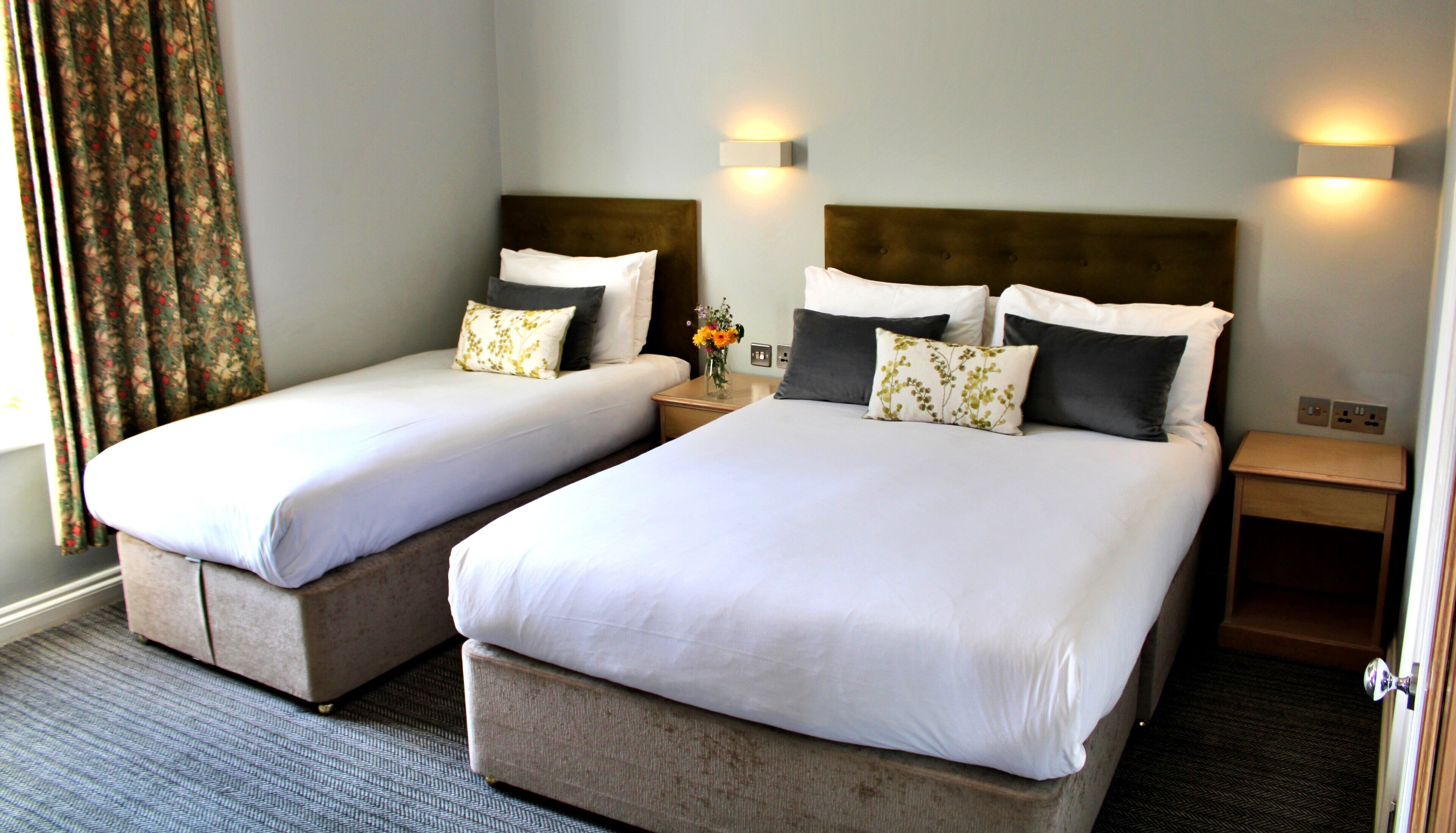 A hotel room with two beds, a nightstand, a vase with flowers, and a window with curtains.