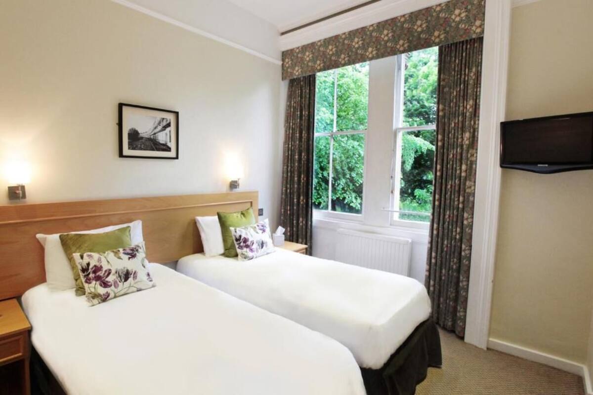 A hotel room with two beds, a television, a window with curtains, and a framed picture on the wall.