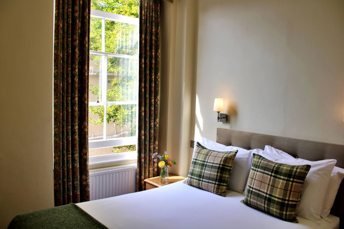 A neatly made bed with white linens and plaid pillows, a bedside table with a vase of flowers, and a window with curtains.
