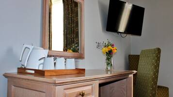 A hotel room with a wooden desk, a mirror, a flat-screen TV, a green upholstered chair, and a vase with flowers.