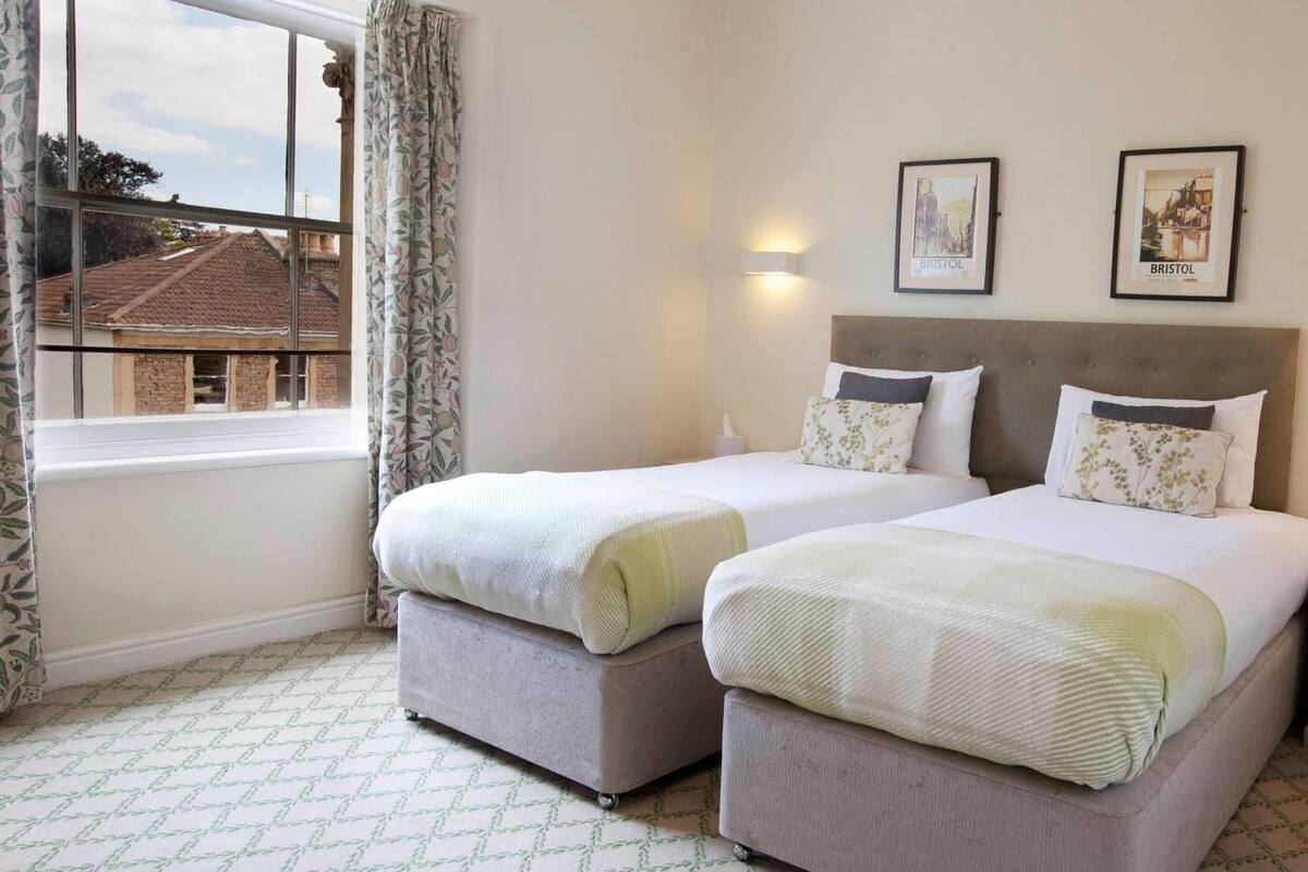 A hotel room with two beds, a window with curtains, a wall-mounted light, and two framed pictures.