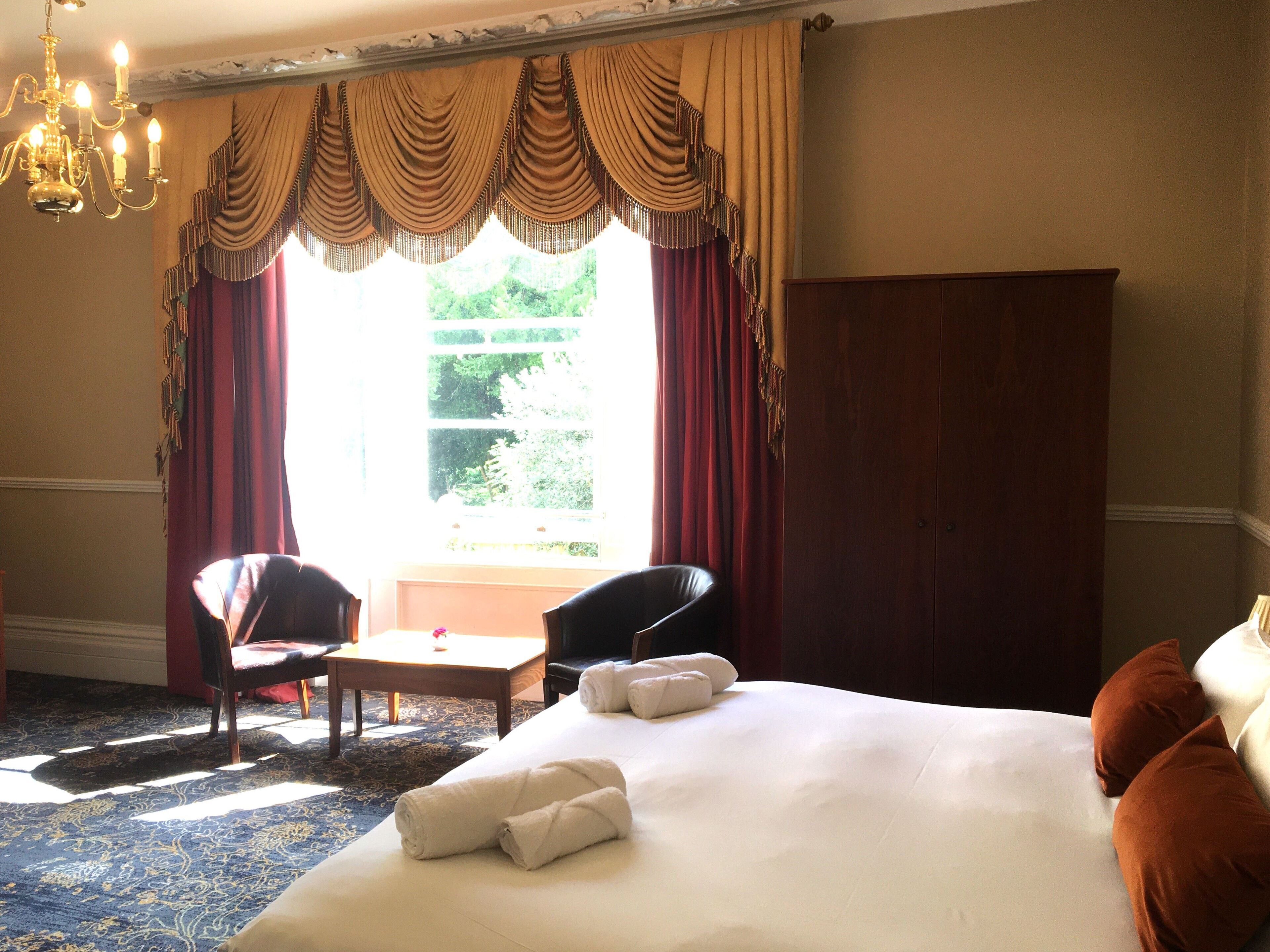 Quarto de hotel com cama grande, duas cadeiras, uma mesinha e um candelabro.
