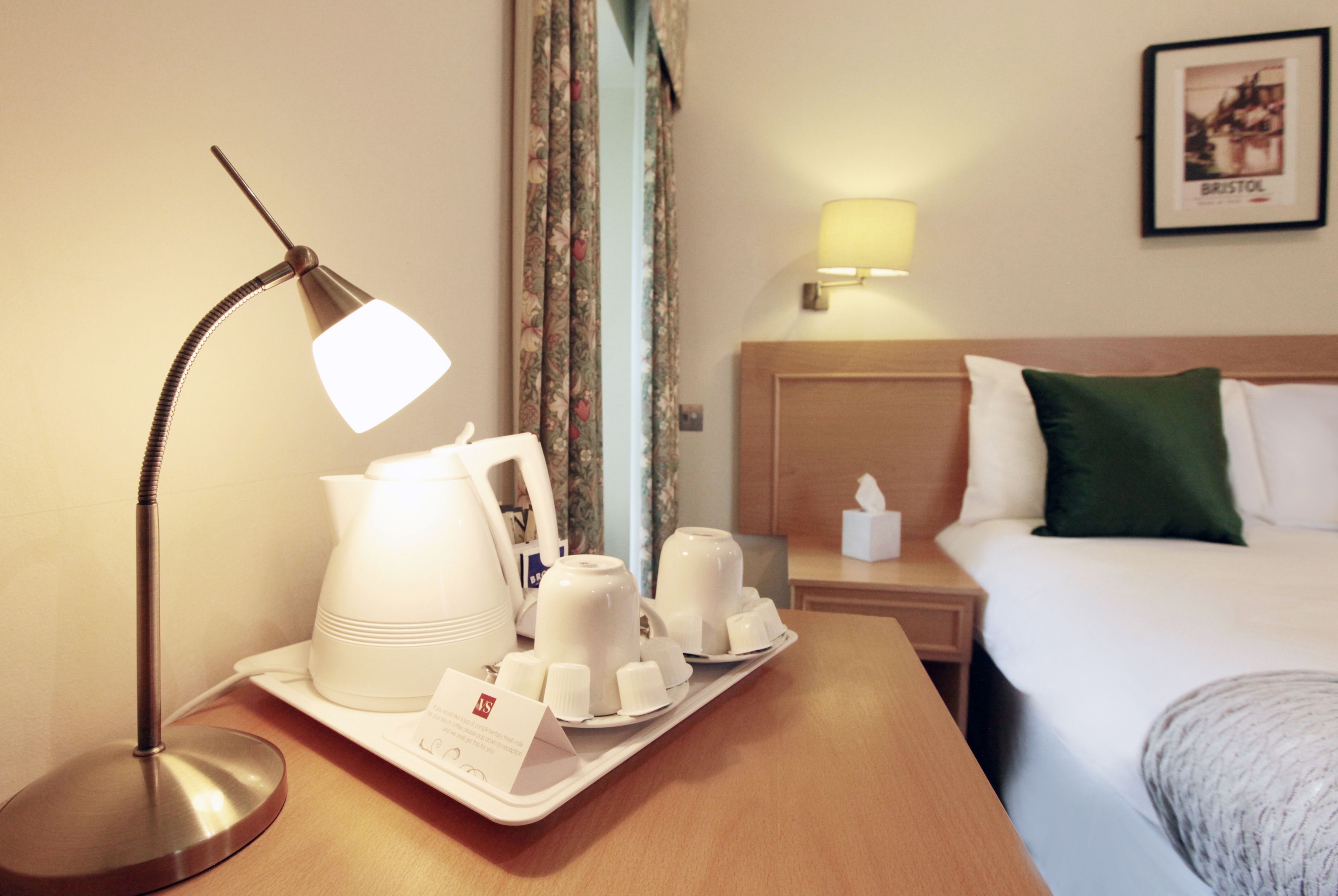 Habitación de hotel con mesita de noche que incluye una lámpara, una bandeja con tazas blancas y un hervidor de agua, una lámpara adicional, un cuadro en la pared y una cama bien hecha con un almohadón verde.