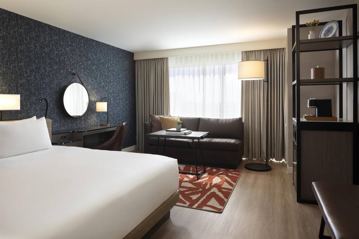 Una habitación de hotel con una cama grande, un escritorio con silla, un sofá, una estantería y un ventanal con cortinas.