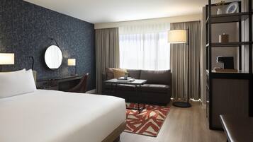 A hotel room with a large bed, a desk with a chair, a sofa, a bookshelf, and a large window with curtains.
